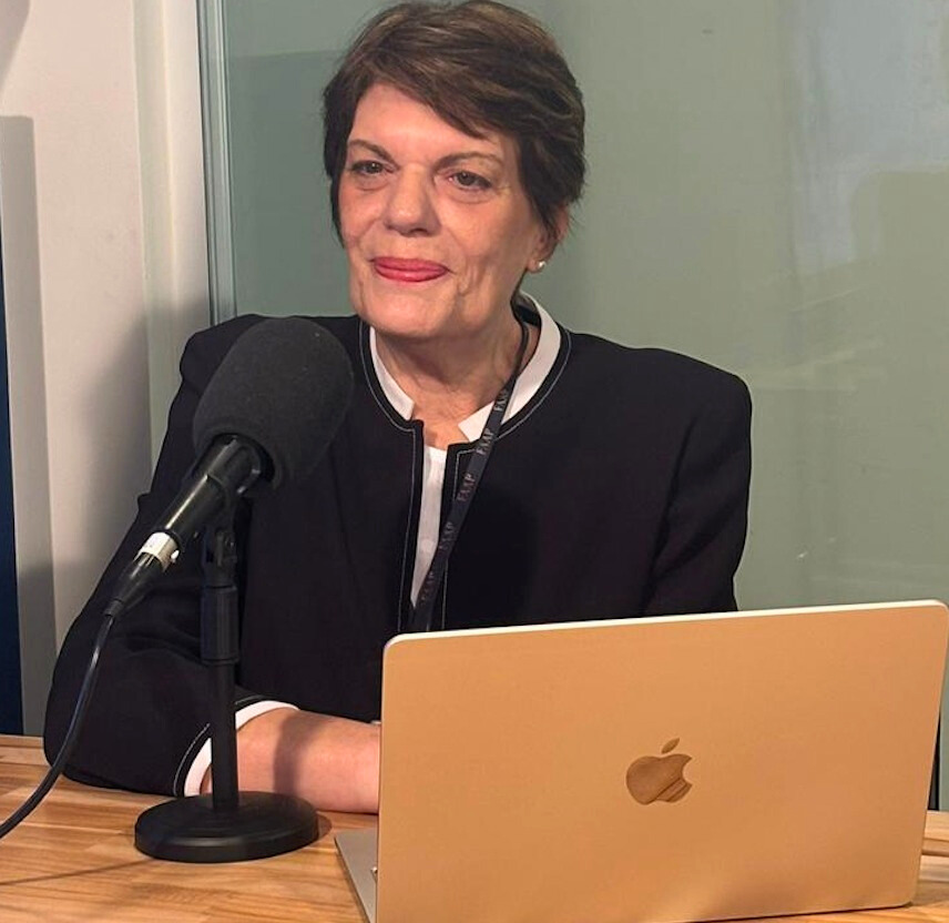 Pessoa sentada em estúdio de gravação, diante de um microfone e de um notebook sobre a mesa.